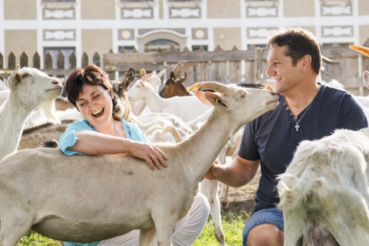 Eine Frau und ein Mann in der Hocke mitten in einer Ziegenherde vor einem Bauernhaus.