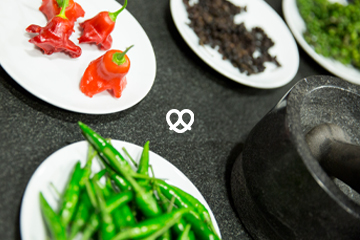 Chilischoten, Paprika und verschiedene Pfeffersorten auf weißen Tellern, daneben steht ein schwarzer Marmor-Mörser mit Stößel.