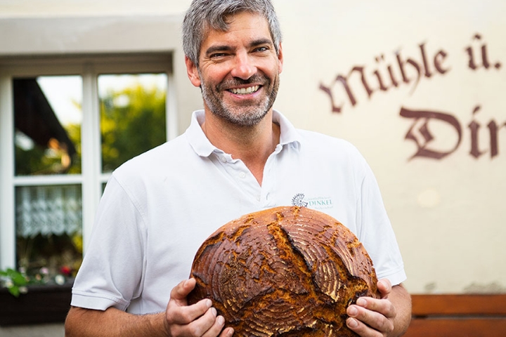 Porträt eines Bäckers vor seiner Bäckerei mit einem runden Brotlaib in den Händen.