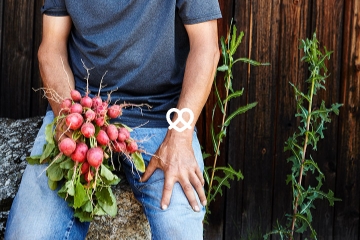 Gärtner sitzt mit einem Bund Radieschen in der Hand auf einem Stein vor Holzwand.