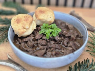 Eine Schüssel steht auf einem Holztisch, umgeben von grünen Zweigen. In der Schüssel ist Rehgulasch, dekoriert mit Petersilie und zwei Gebäckstücken, den Hauberlingen.
