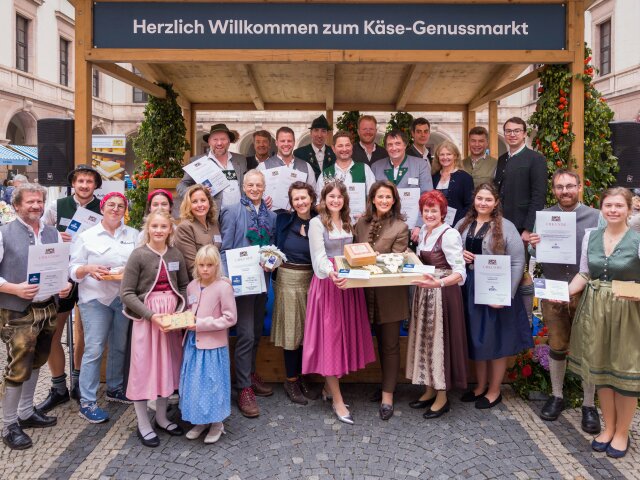 Gruppenfoto der Gewinner des Käsewettbewerbs 2025: eine Gruppe von Menschen in bayerischer Tracht, einige halten Urkunden, einige halten Holzbretter mit Käse